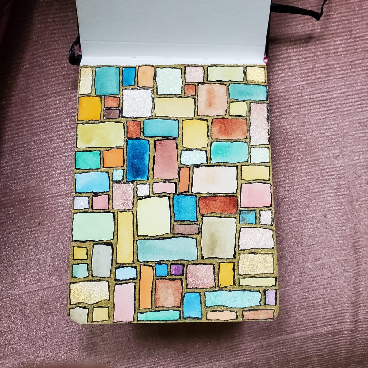 A watercolor journal page of uneven boxes in varying sizes and colors outlined in black ink against a gold acrylic marker background. The journal sits on a textured mauve chair cushion.